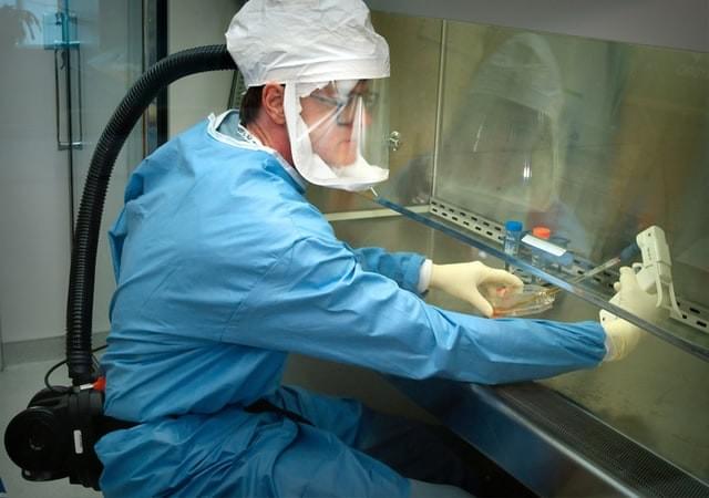 An image of a medical researcher in a blue suit with gloves, a hood, a ventilator, and clear face mask working with a pipette