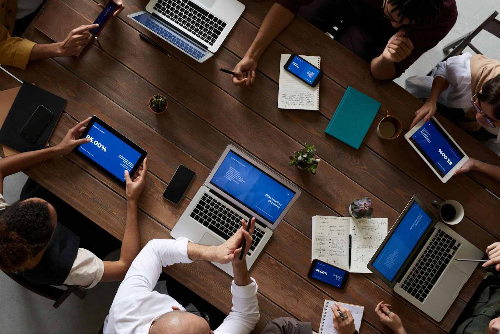 A group of people gathered around a table viewed from above using laptops, tablets, smartphones, and paper documents to work together